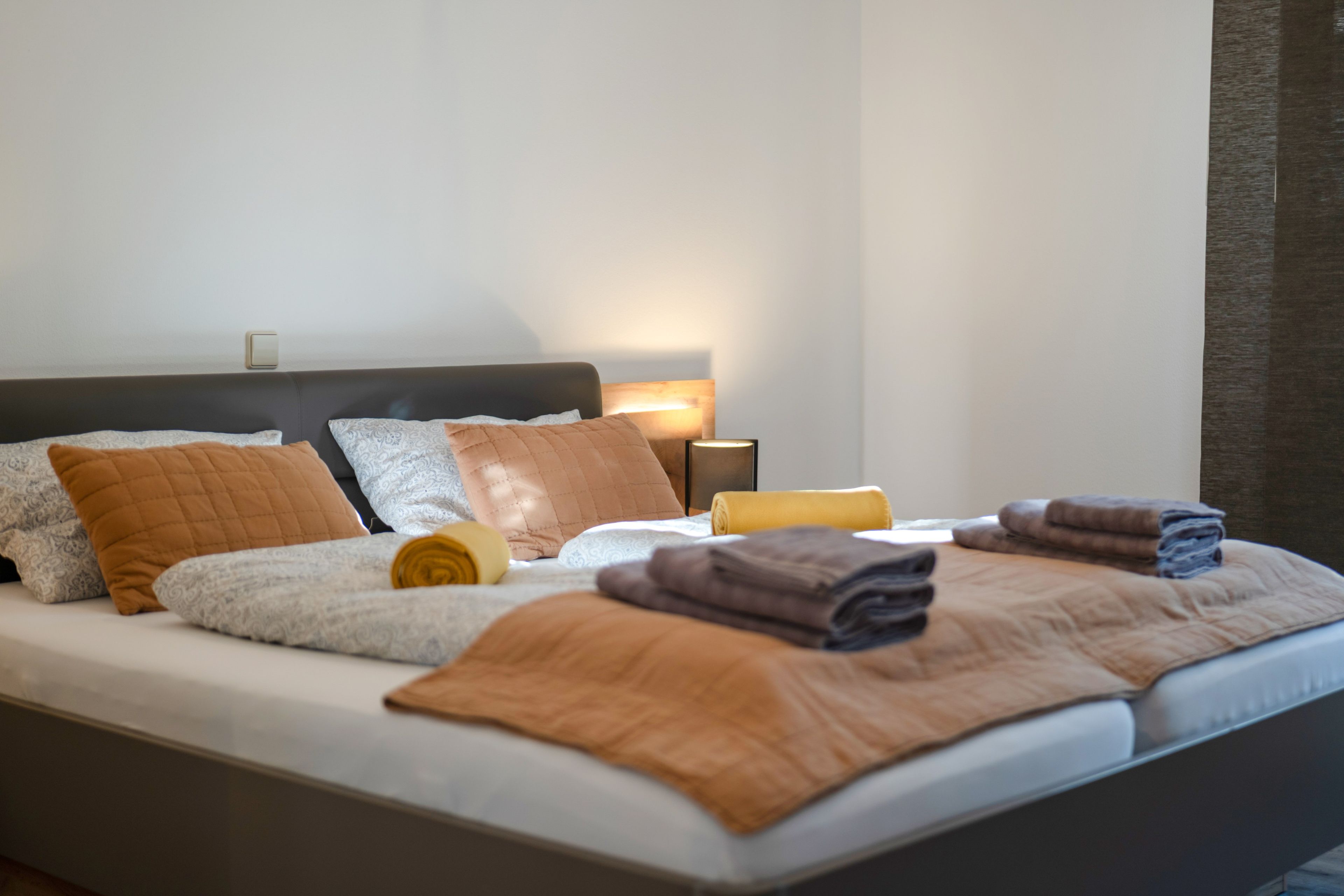 A neatly made bed with pillows and a blanket, a bedside table with a lamp, and a wall switch.