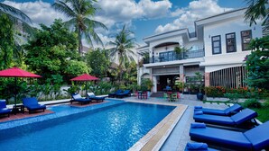 Une piscine entourée de chaises longues bleues et de parasols rouges, le tout bordé d’une végétation luxuriante, avec un bâtiment à deux étages doté d’un balcon.