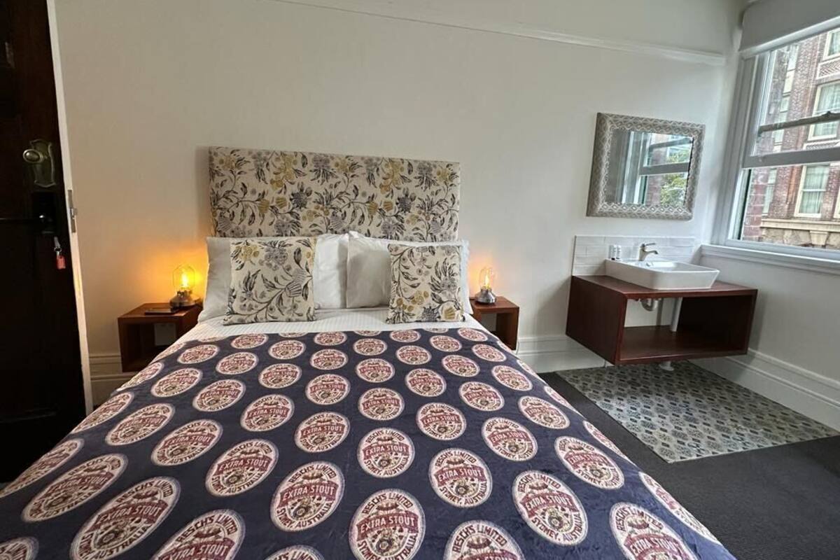 Habitación de hotel con cama, mesita de noche, espejo, ventana y baño con lavabo y espejo.