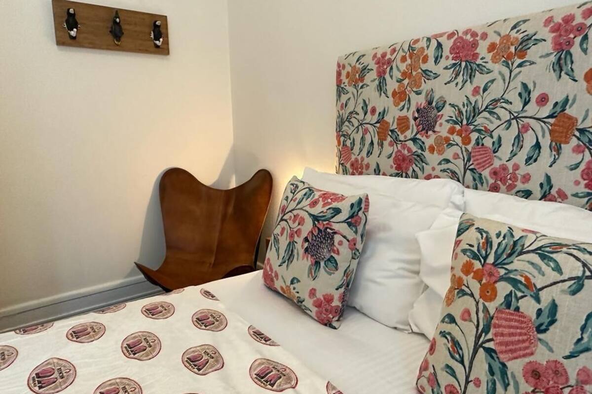 Una cama con almohadas estampadas con motivos florales y un cabecero, una mesita de noche de madera y un estante montado en la pared con tres objetos decorativos.