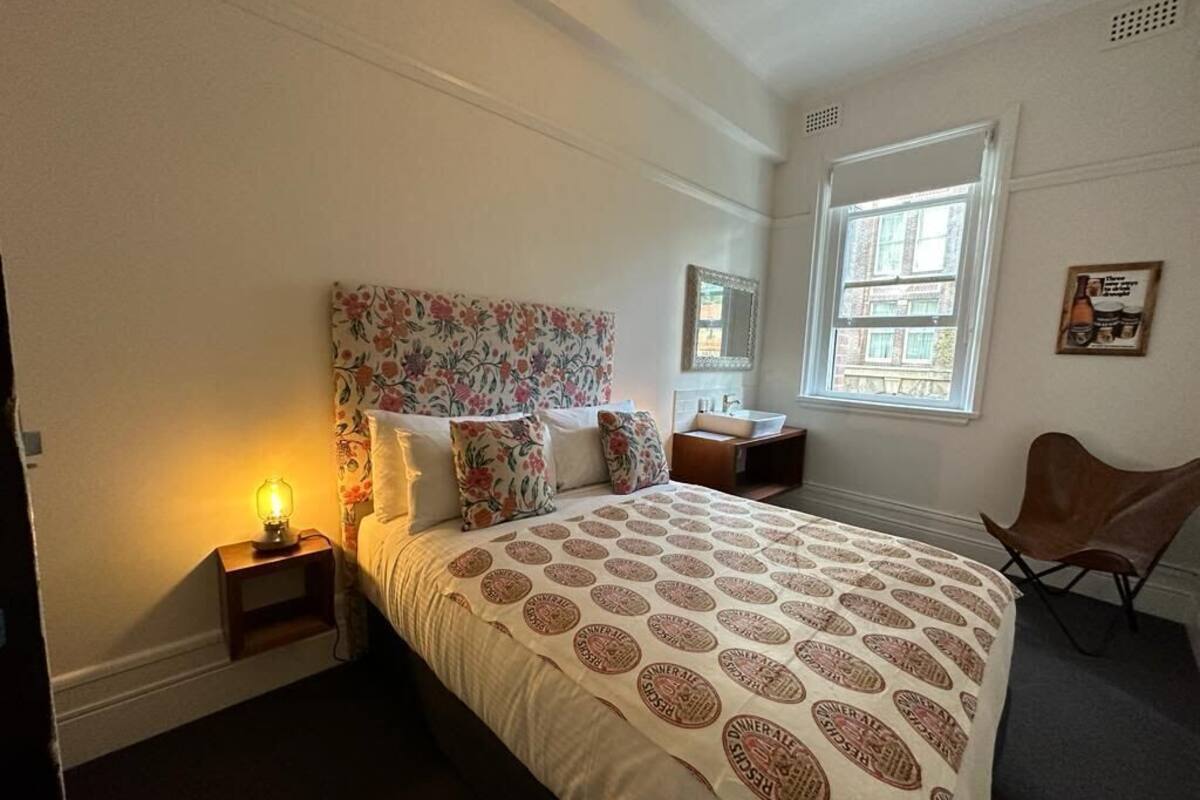 Un dormitorio con cabecera de flores, cama con colcha estampada, mesita de noche con lámpara, una silla y ventana con vista a edificios.