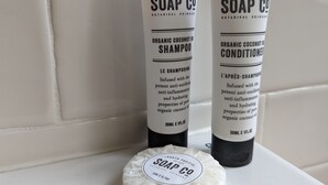 Deux bouteilles de shampooing et d’après-shampooing à l’huile de coco biologique de la marque South Pacific Soap Co., ainsi qu’un pain de savon, le tout posé sur une surface blanche.