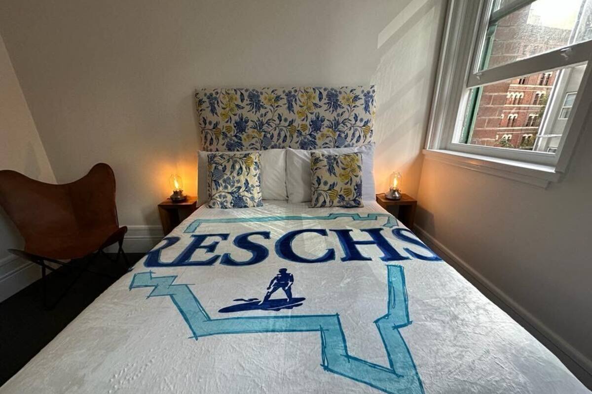 Un dormitorio con una cama que presenta un nombre grande bordado y un logotipo, un cabecero estampado, dos mesitas de noche con lámparas, y vistas a edificios a través de una ventana.