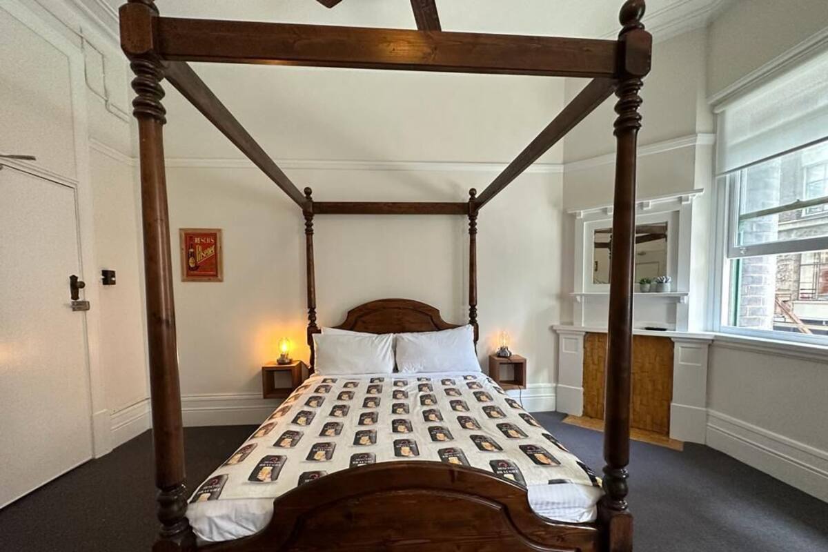 Una cama con dosel con una colcha estampada, una mesita de noche con una lámpara, y una ventana con vistas al exterior.