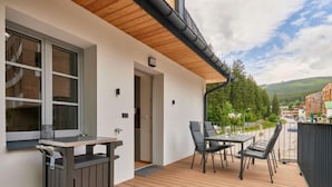Een balkon met een houten terras, een tafel en stoelen, en een uitzicht op een stad en heuvels op de achtergrond.