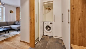 Een modern appartement met een grijze bank, houten vloeren, een wasmachine en een houten garderobe.