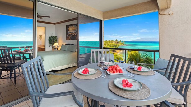 A balcony with a view of the ocean, a dining table set with plates and glasses, and a bed with a green bedspread. - MILLION DOLLAR VIEW!!  2 BDRM OCEANFRONT CONDO WEST MAUI. NEW AC IN EVERY ROOM. (Lahaina)