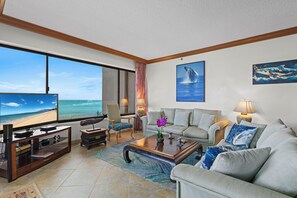 A living room with a large TV, comfortable seating, a coffee table, and a view of the ocean. - MILLION DOLLAR VIEW!!  2 BDRM OCEANFRONT CONDO WEST MAUI. NEW AC IN EVERY ROOM. (Lahaina)