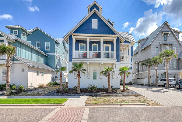 A two-story residential building with a blue exterior, a balcony with white railings, and palm trees in the front yard. - All-new waterfront home with on-site pool, golf, & easy beach access (Port Aransas)