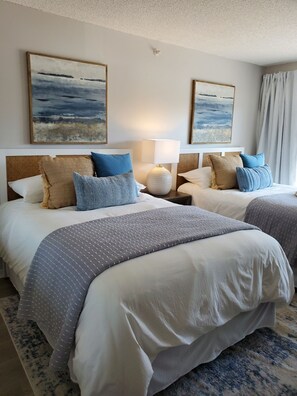 Two large beds with pillows and throw blankets, a bedside table with a lamp, and abstract artwork adorn this inviting space.