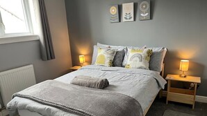 Una cama bien hecha con edredón gris y blanco, dos mesitas de noche con lámparas, y tres cuadros en la pared sobre la cama.