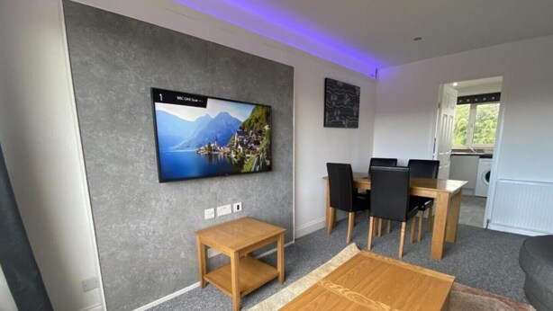 Een moderne woonkamer met een flatscreen televisie aan de muur, een houten salontafel, een eethoek met een tafel en stoelen, en een keuken zichtbaar door een open deur.