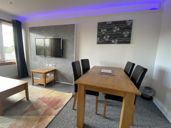 Una sala moderna con una mesa de comedor de madera, sillas negras, un televisor de pantalla plana montado en la pared y una alfombra estampada en el suelo.