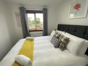 Una cama bien tendida con ropa de cama blanca y amarilla, cortinas grises y un cuadro en la pared.
