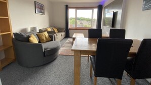 Een moderne woonkamer met een grijze bank, een houten salontafel en zwarte stoelen.
