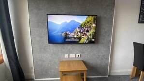 Een moderne woonkamer met een flatscreen televisie aan een grijze muur, een klein houten bijzettafeltje en een houten salontafel.