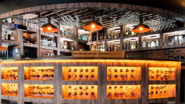 Zona de bar con estanterías de madera mostrando botellas, una barra central con repisas iluminadas y lámparas colgantes.