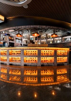 Un espace bar avec des étagères en bois où sont exposées des bouteilles, un comptoir central avec des étagères éclairées et des lampes suspendues.