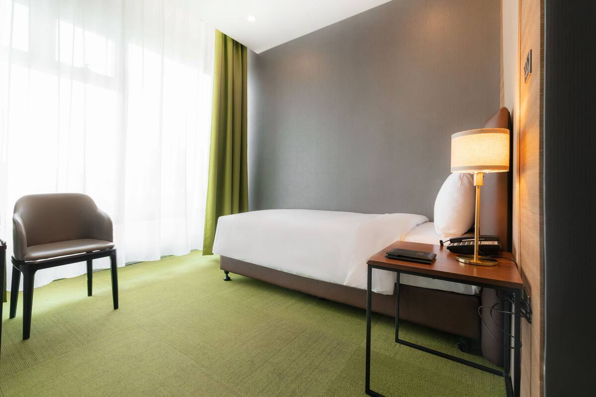 Une chambre d’hôtel avec un lit, une chaise, un bureau avec une lampe et une fenêtre avec des rideaux légers.