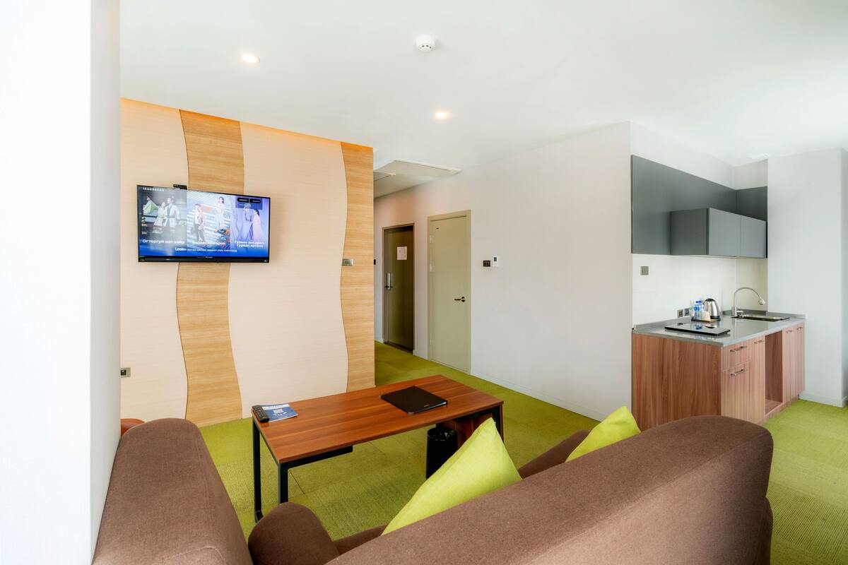 Une chambre d’hôtel moderne équipée d’un canapé, d’une petite table, d’un téléviseur fixé au mur, d’une kitchenette et d’une porte donnant sur une autre pièce.