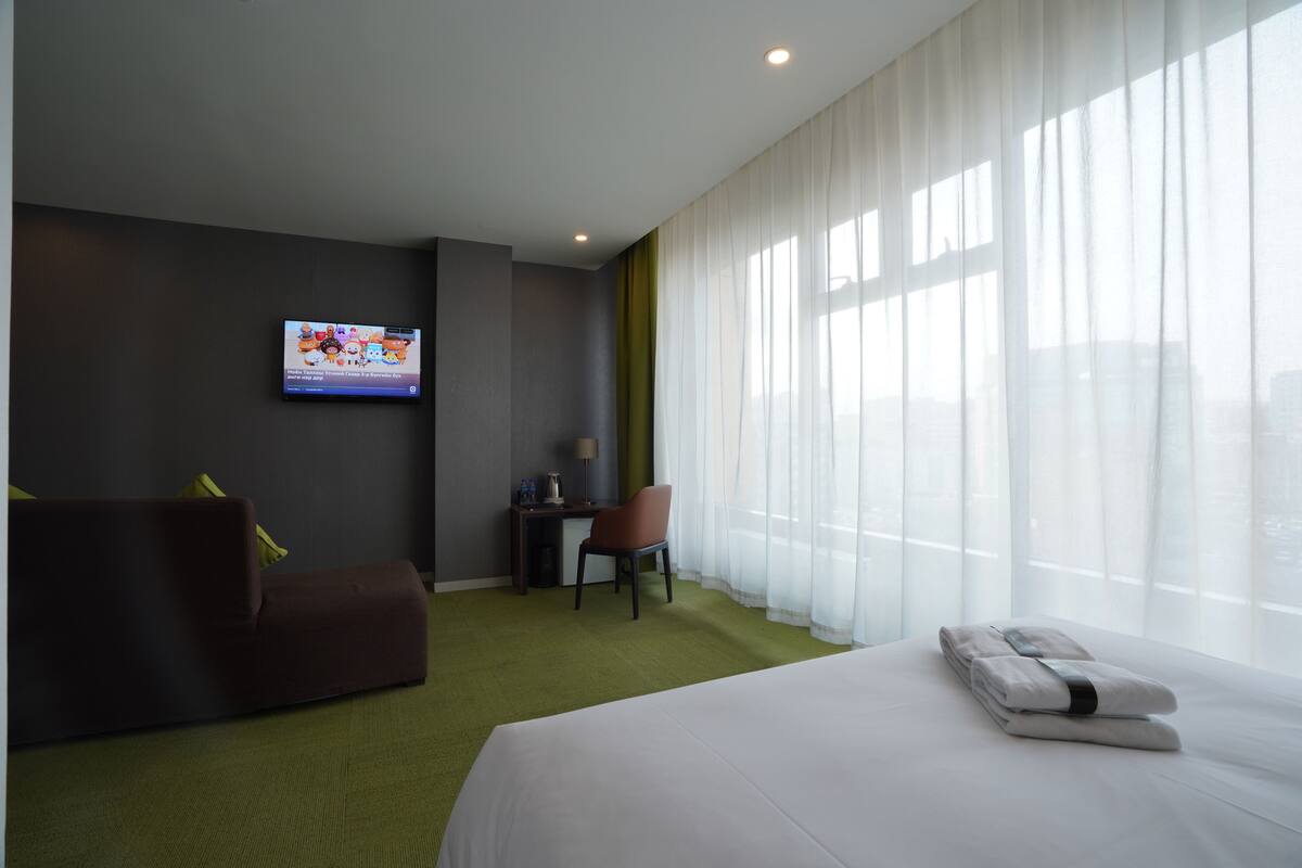 Une chambre d’hôtel équipée d’un lit, d’une télévision, d’un bureau, d’une chaise et d’une fenêtre avec des rideaux légers.