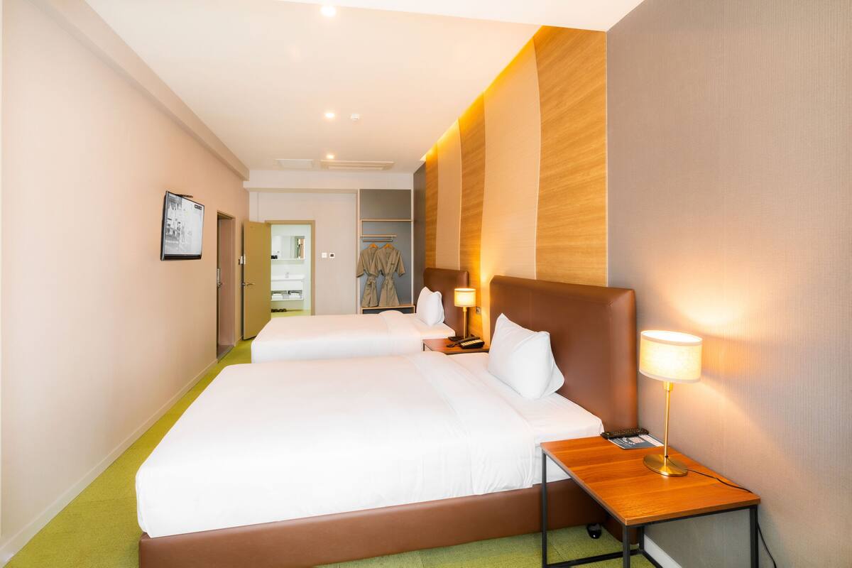 Une chambre d’hôtel avec deux lits, une tête de lit en bois, une table de chevet avec une lampe et une moquette verte.