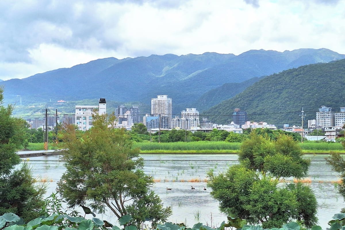 Un paisaje urbano con un río al frente, rodeado de vegetación y montañas al fondo.