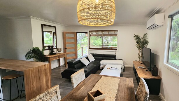 A modern living room with a sofa, coffee table, and dining table. There is a large window with a view of greenery outside. - Tallowood Treehouse  (East Ballina)