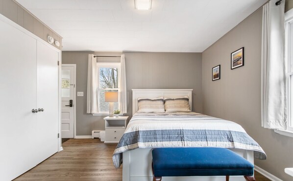 A bedroom with a bed, a nightstand, a window with curtains, a door, and a wall with two framed pictures.