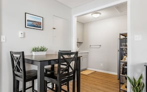 A modern kitchen with white cabinets, a wooden dining table, and chairs. There is a potted plant on the table and a view of the outside through the window.