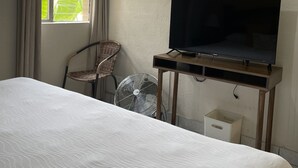 A hotel room with a bed, a television on a wooden stand, a wicker chair, a standing fan, and a window with curtains. - Inari Guatemala (Guatemala City)