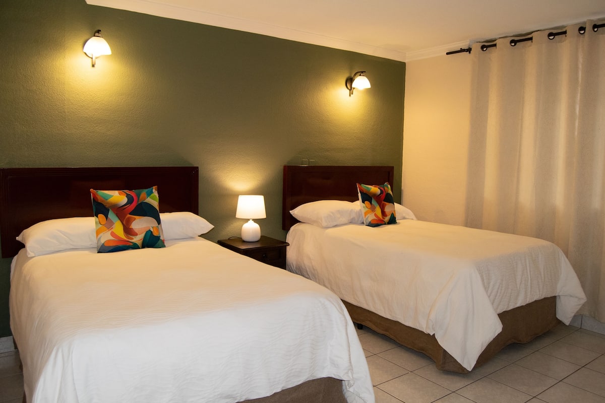Deux lits simples avec du linge de lit blanc, une table de chevet avec une lampe et un luminaire fixé au mur.