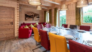 Uma sala de jantar com decoração em madeira, uma mesa grande posta para seis pessoas e um sofá com almofadas coloridas.