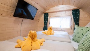 Quarto de cabana aconchegante com paredes de madeira, uma TV de tela plana na parede, duas camas com toalhas amarelas e uma janela com cortinas.