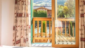 A room with a large sliding glass door leading to a balcony with a wooden railing, a view of a mountain landscape, and patterned curtains.