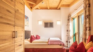 A bedroom with a wooden wardrobe, a bed with red bedding, a red sofa, and a window with patterned curtains.