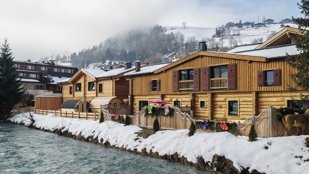 A wooden lodge with a river in front, featuring a balcony with red shutters and a view of a snowy hillside.