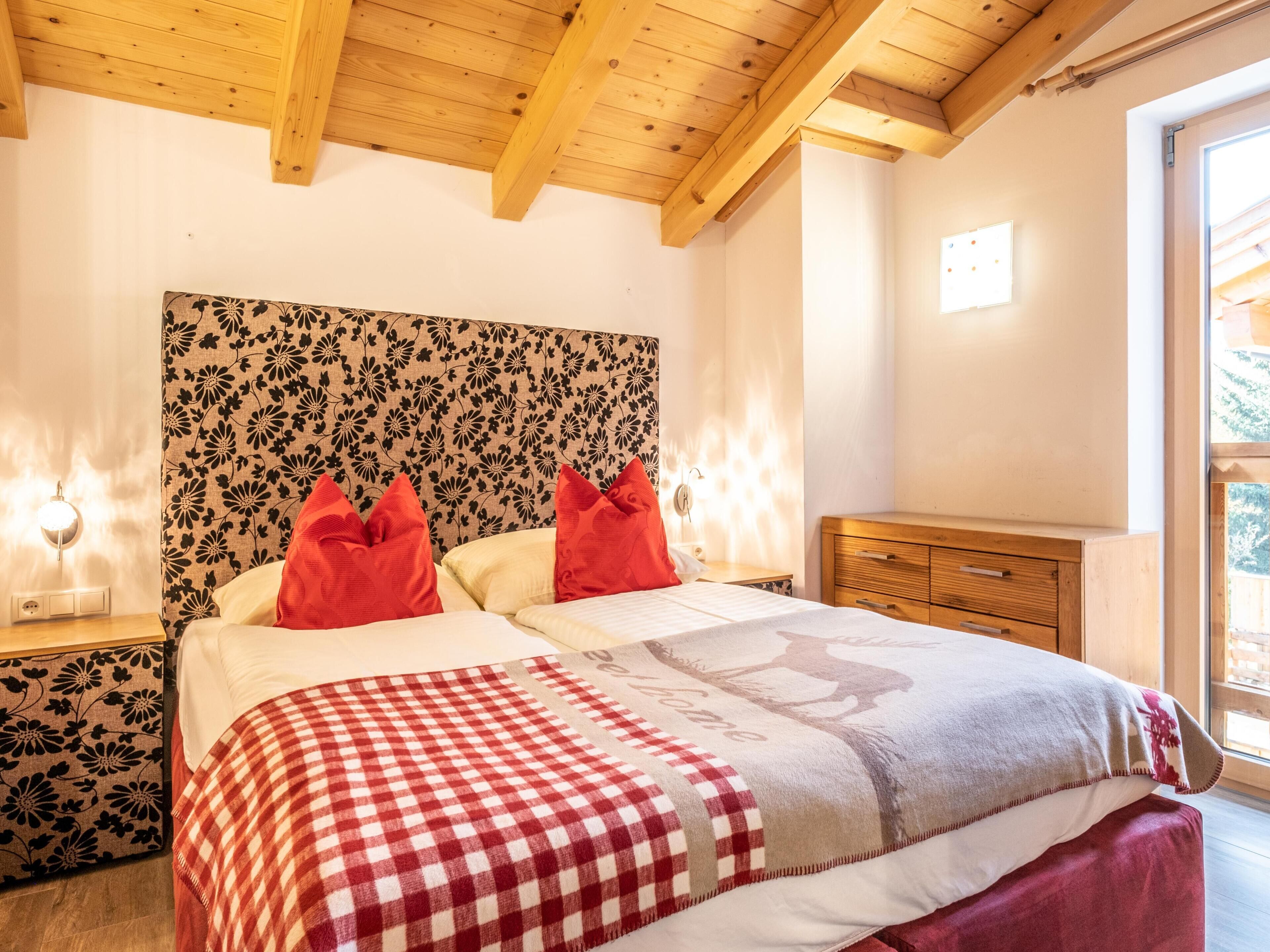 Ein Schlafzimmer mit Holzbalkendecke, einem blumengemusterten Kopfteil, einem Bett mit rot-weißer Bettwäsche und einem Nachttisch mit Lampe.