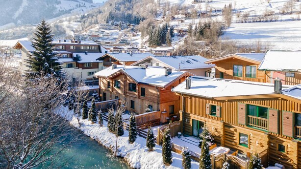 A winter resort with wooden cabins, a river, and snow-covered hills. - Group house with sauna in the heart of Kaprun (Kaprun)