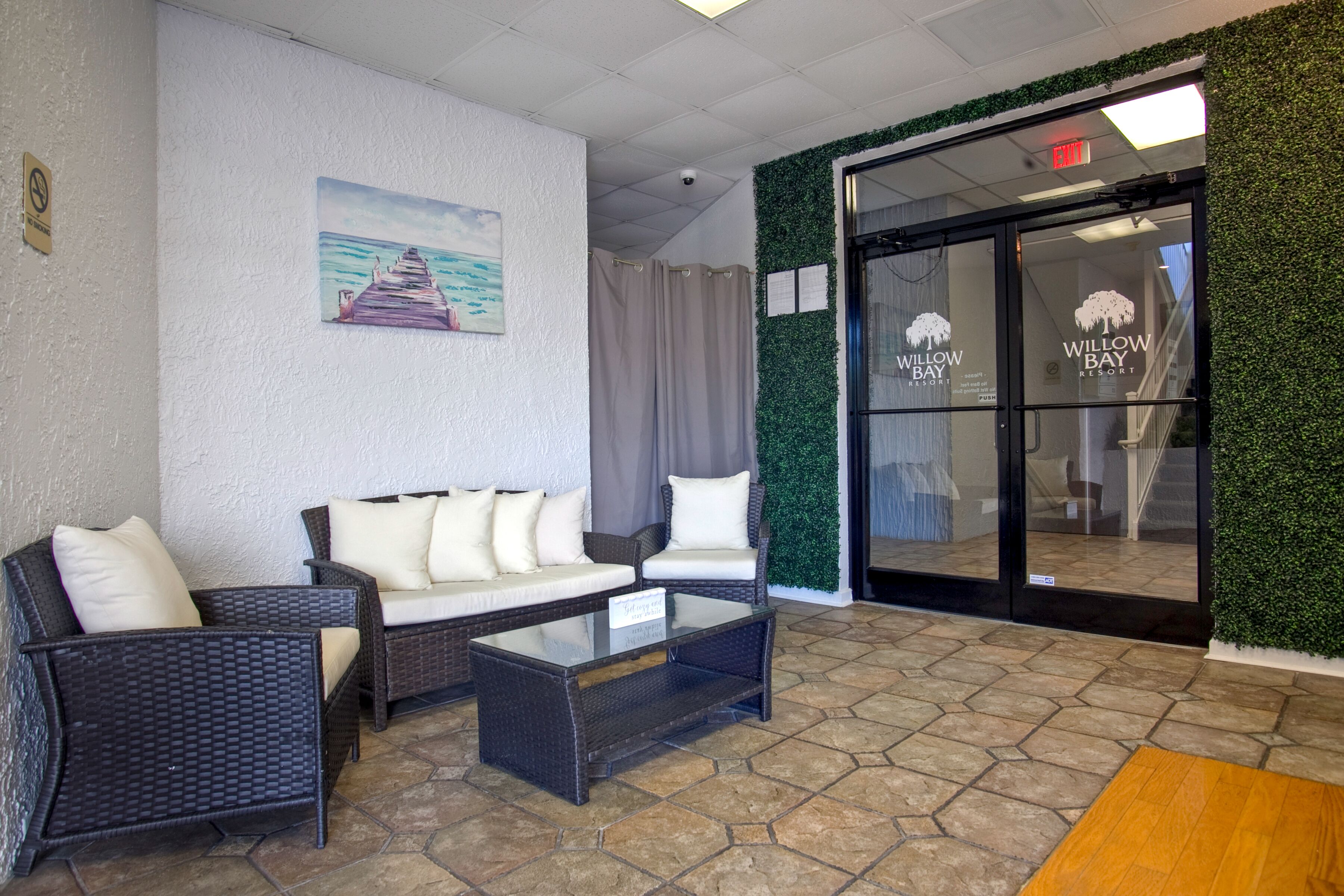 A lobby area with a white textured wall, a painting of a pier, wicker furniture, and a glass door leading to the entrance of Willow Bay Resort.