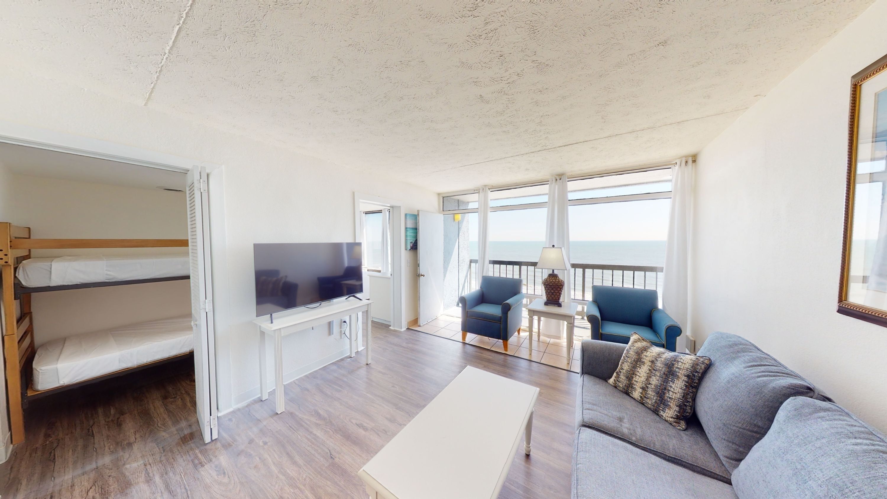 A room with a bunk bed, a flat-screen TV, a sofa, two armchairs, a small table, and a view of the sea through large windows.