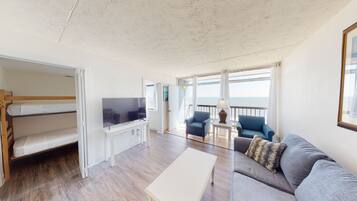 A room with a bunk bed, a flat-screen TV, a sofa, two armchairs, a small table, and a view of the sea through large windows.