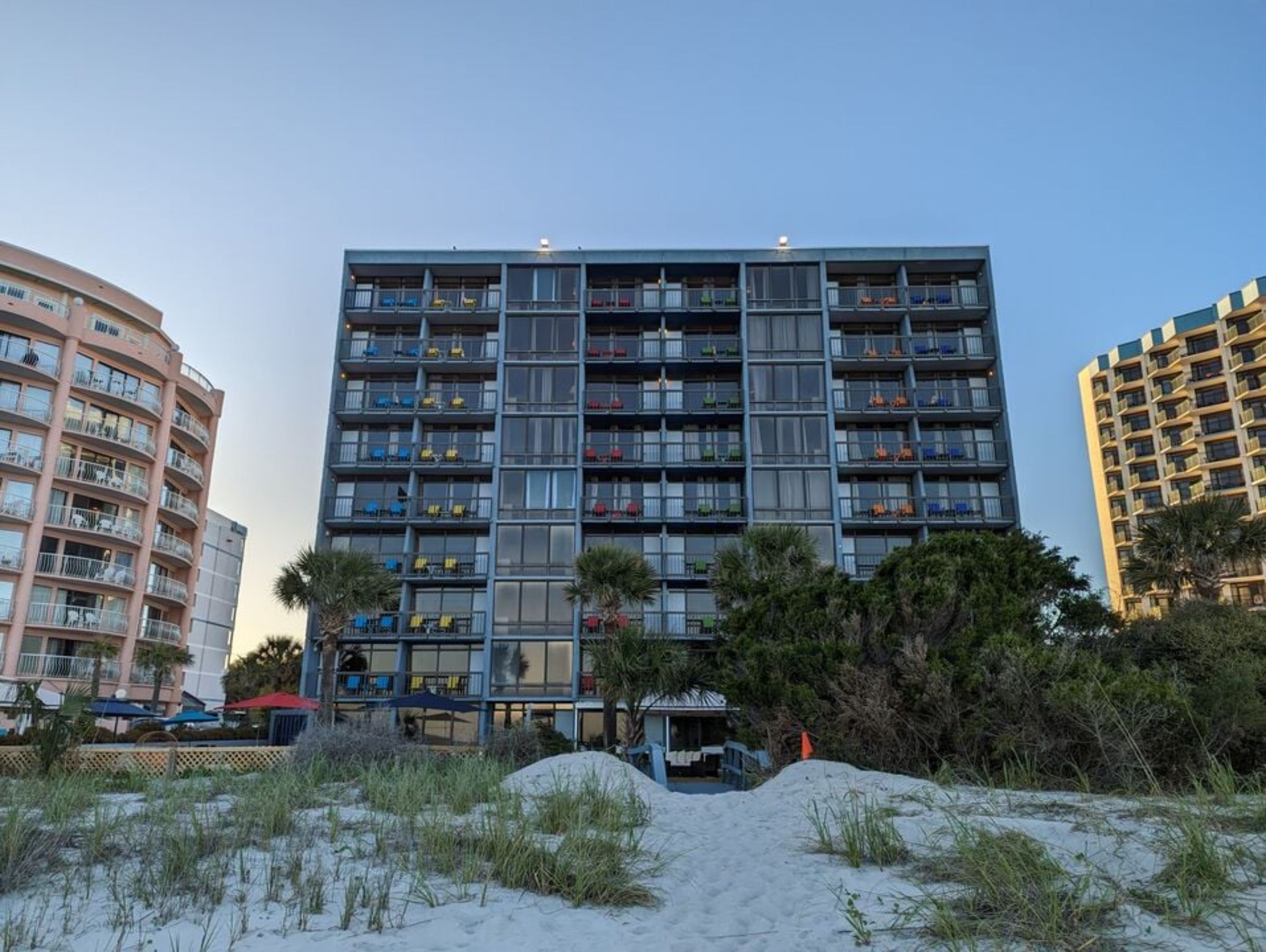 Un hotel de varios pisos con balcones, situado cerca de una playa con dunas de arena.