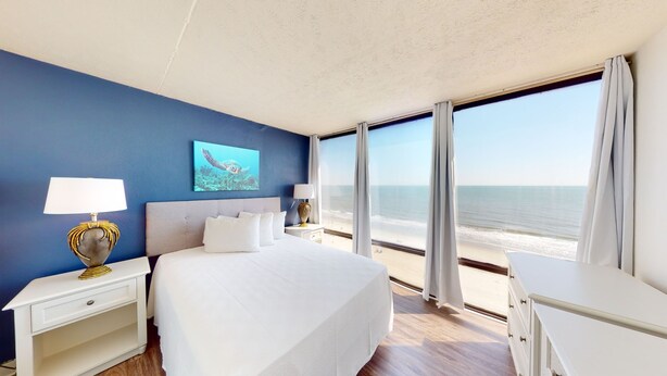 A hotel room with a large bed, two bedside tables with lamps, a painting of a sea turtle, and a view of the beach. - Willow Bay Resort  (Myrtle Beach)