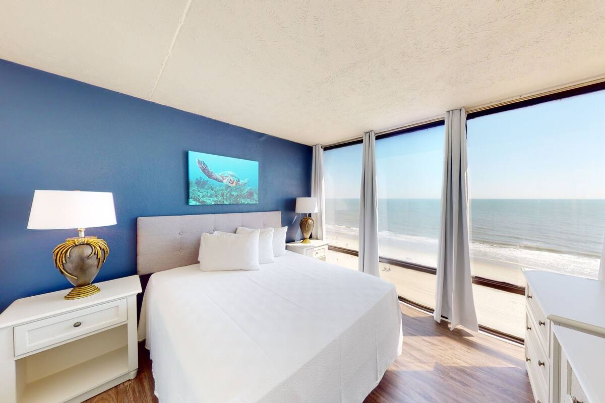 Ein Hotelzimmer mit einem großen Bett, zwei Nachttischen inklusive Lampen, einem Bild von einer Meeresschildkröte und Blick auf den Strand.