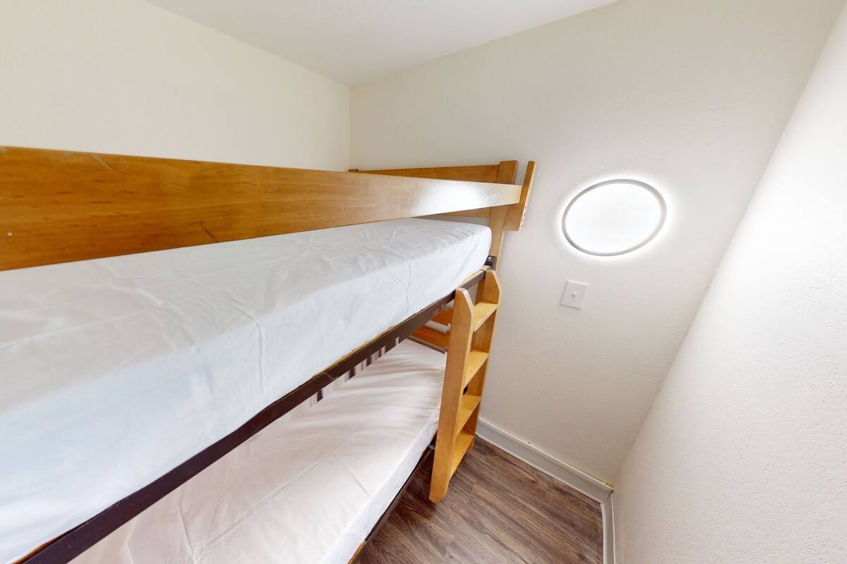 Ein Stockbett mit Holzrahmen und weißer Bettwäsche in einem Zimmer mit weißen Wänden und Holzboden.