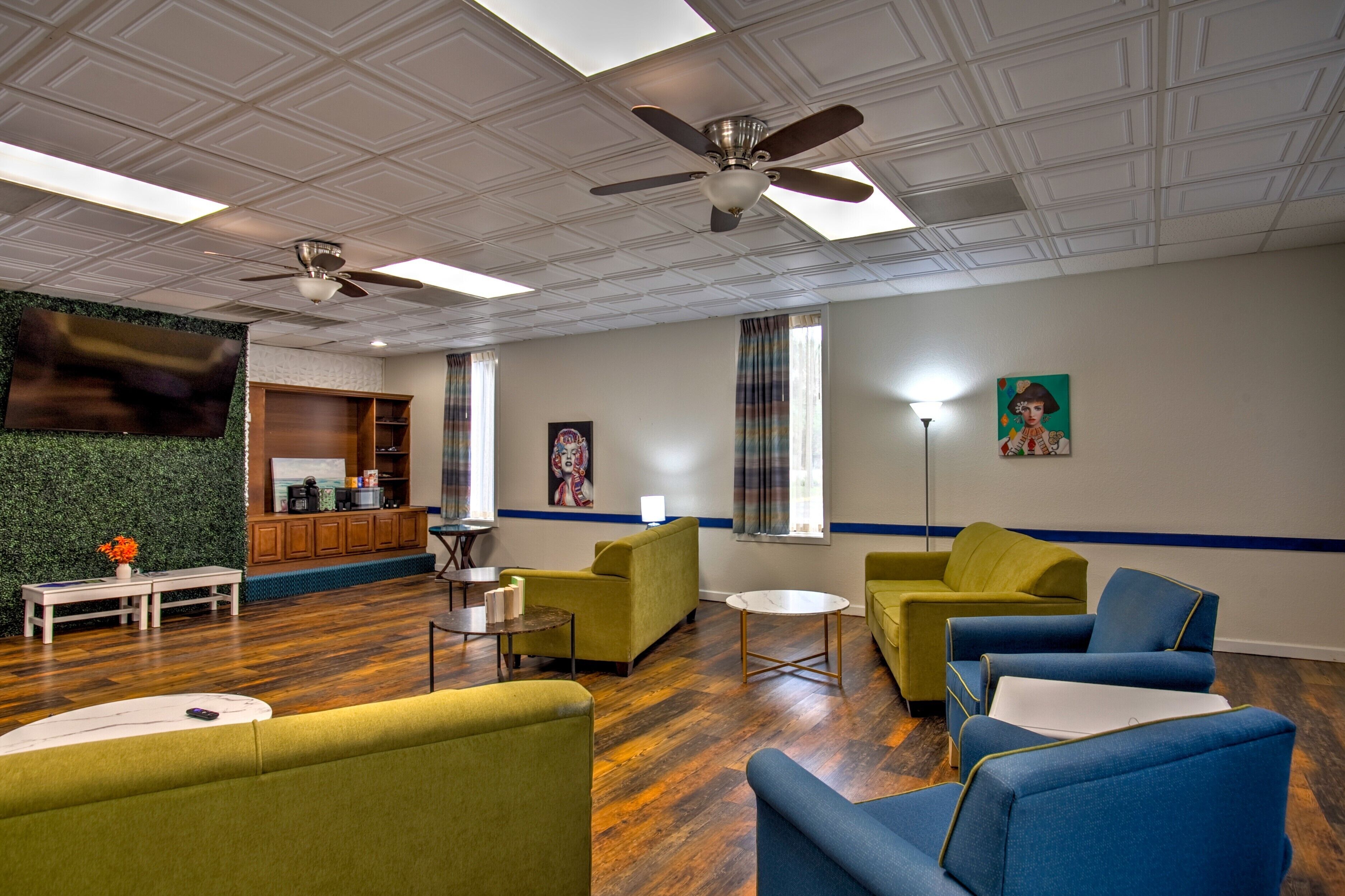 A spacious living area with a wooden floor, green and blue sofas, a ceiling fan, and a flat-screen TV mounted on a green wall.