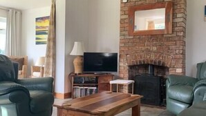 Een woonkamer met een open haard van baksteen, groene leren banken, een houten salontafel en een televisie.