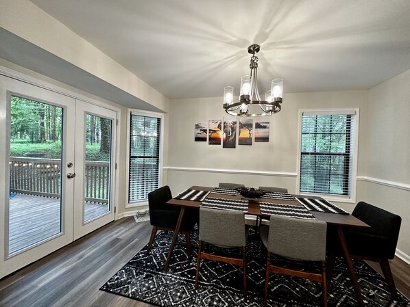 A dining area with a chandelier, a table with chairs, and a view of the outdoors through large windows. - Downtown Boho Oasis w/ Firepit & Game Rm w/ Marvel Arcade, Sleeps 8, Wills Park (Alpharetta)