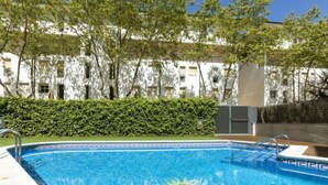 Ein Schwimmbecken mit Sonnendesign auf dem Boden, umgeben von einer Hecke und Bäumen, mit einem Gebäude im Hintergrund.